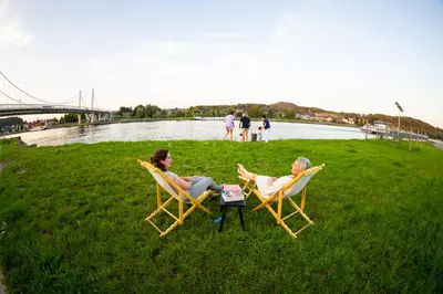 two women sitting in chairs on the grass by a river at De Koperen Geit in Kanne